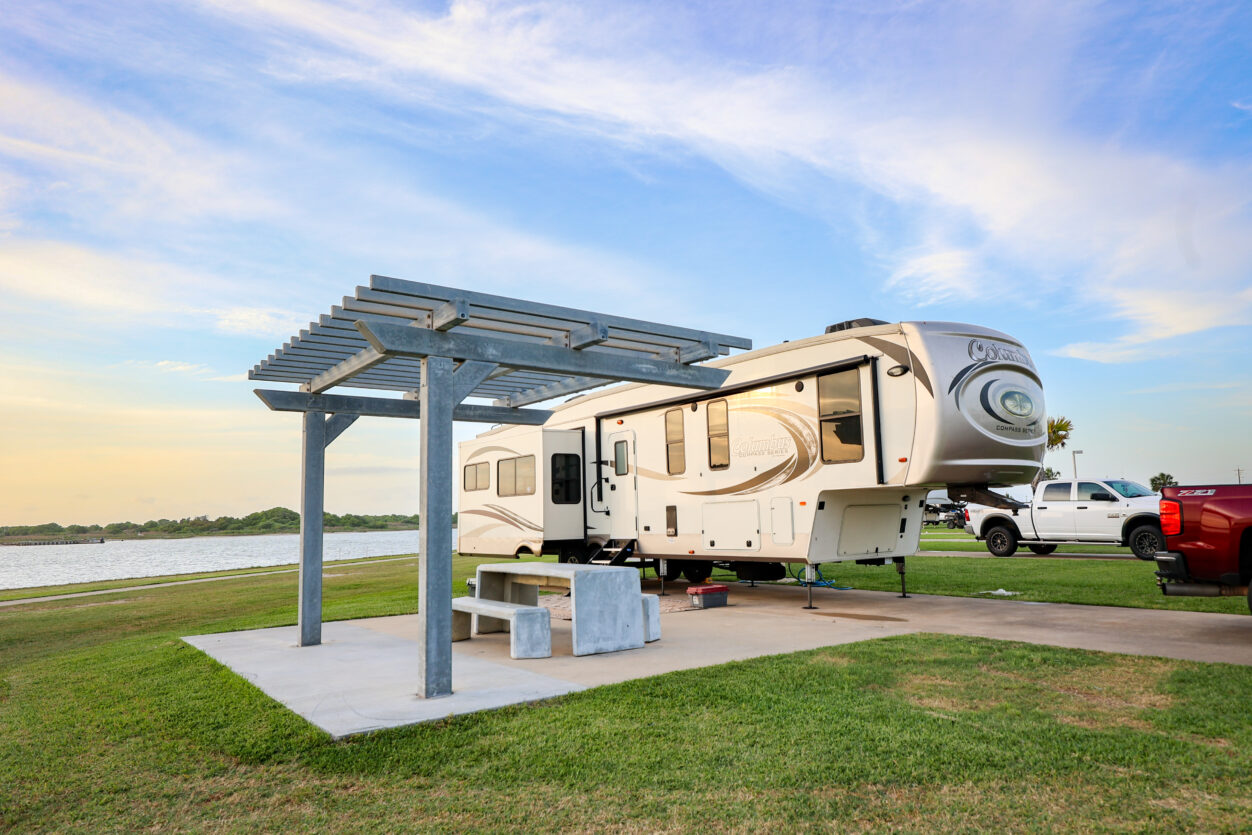 A large RV is parked next to a red pickup truck on a grassy site by a lake. The site has a concrete patio with a metal pergola, picnic table, and benches under a partly cloudy sky at sunset.