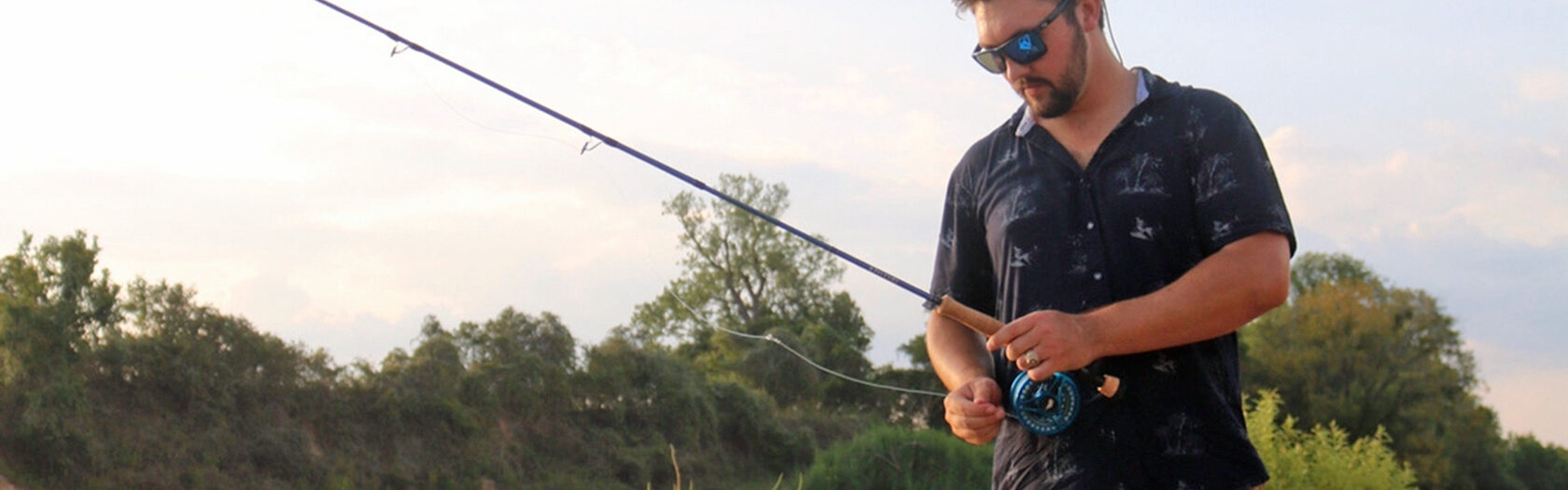 A man wearing sunglasses and a dark shirt stands by a river, holding a fishing rod and adjusting the reel, with trees and shrubs in the background.