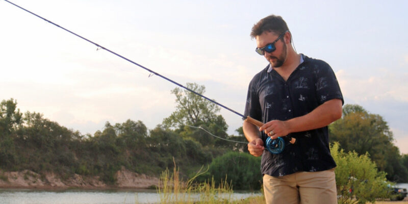 A man wearing sunglasses and a dark shirt stands by a river, holding a fishing rod and adjusting the reel, with trees and shrubs in the background.