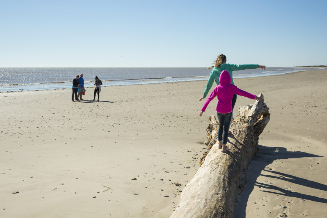Two children in winter attire playing on a beach while people looking for shells in the background.