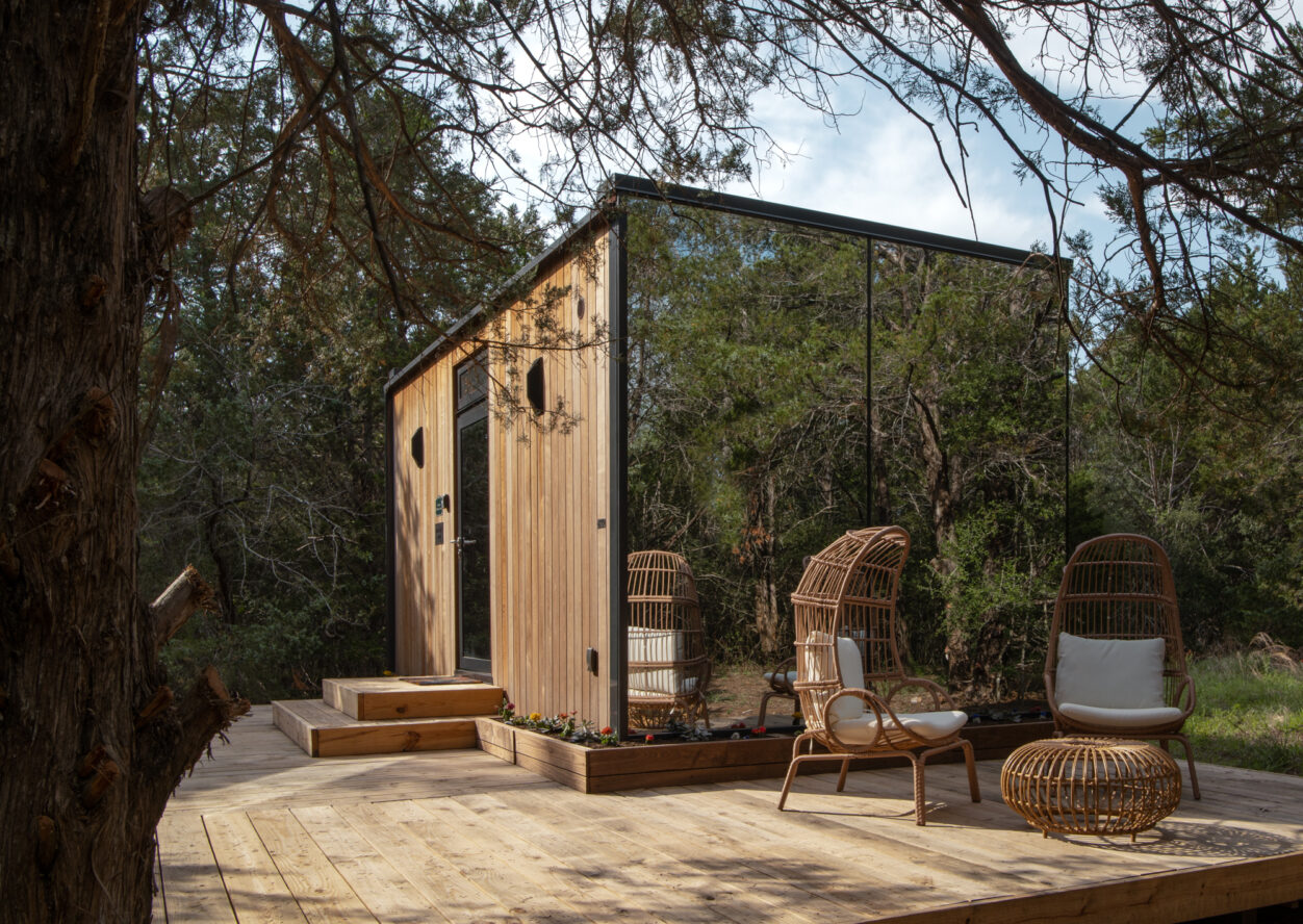 A modern tiny house with wooden siding and large glass windows stands on a wooden deck in a forest. Two wicker chairs, a small table, and potted plants create a cozy outdoor seating area surrounded by trees.