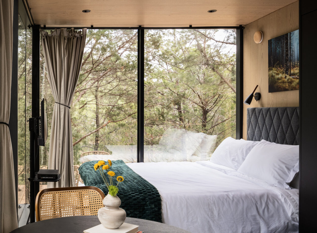 A cozy bedroom with large floor-to-ceiling windows showing a forest view, a neatly made bed with white linens, a round table with a vase of yellow flowers, and light-colored curtains partially pulled aside.