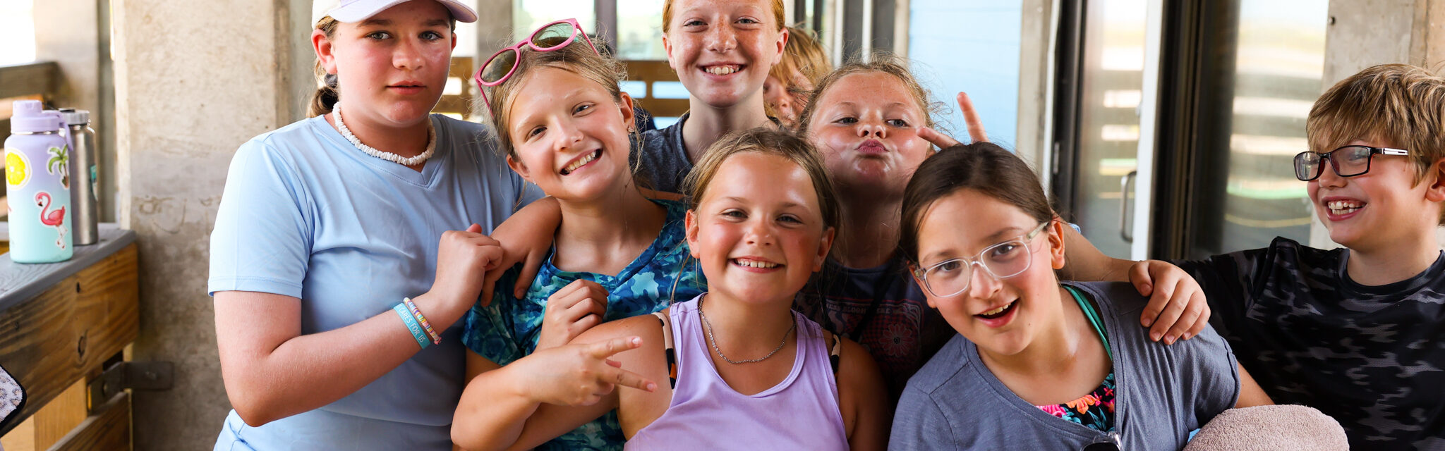 A group of smiling kids pose together on a porch, some making peace signs. They wear casual summer clothes and look happy, with sunlight streaming in from outside.