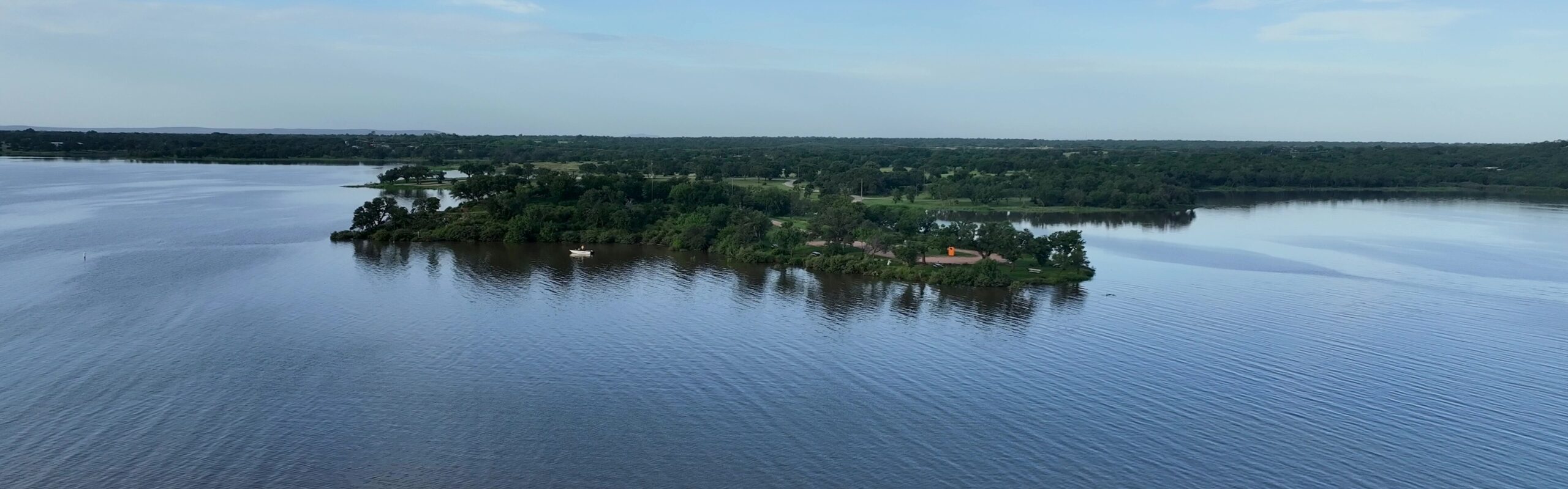 Aerial view of a green, tree landscape with open blue lake water, and distant rolling hills under a blue sky.