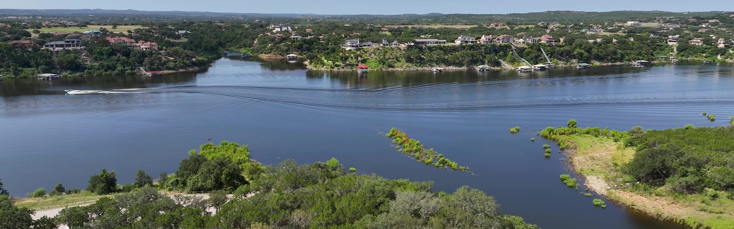 An aerial view of a blue lake with a boat traveling down it and a park with green treetops under a bright blue sky. Low hills and a few buildings are visible in the distance.