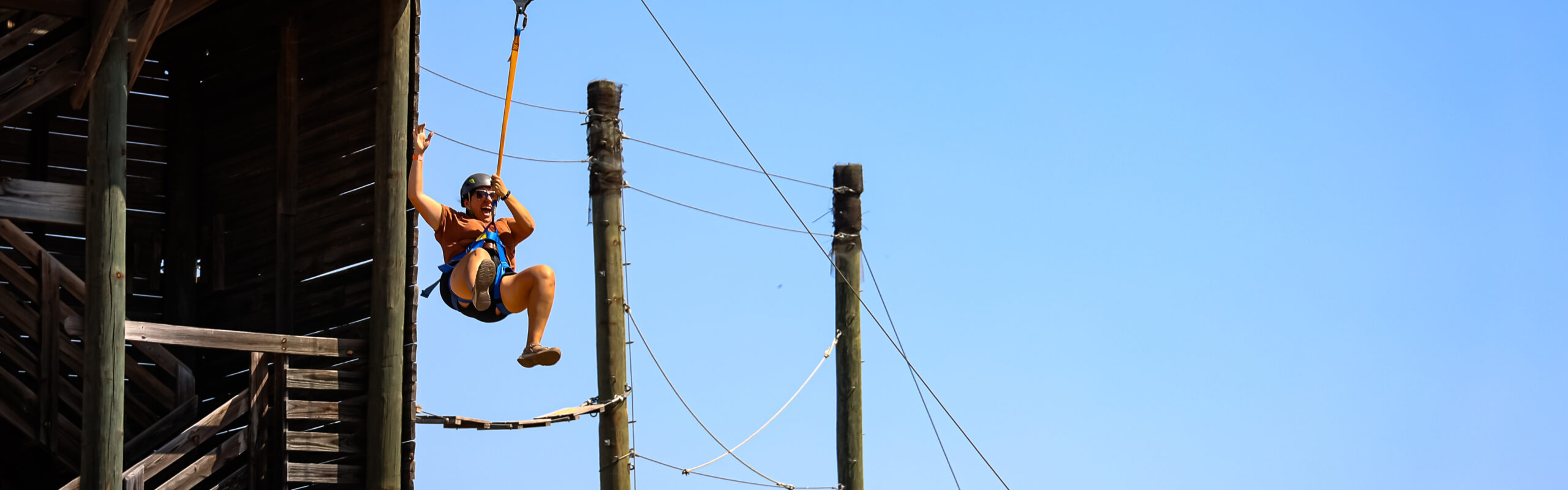 A person wearing a helmet and harness descends from a wooden platform on a zip line against a clear blue sky, with several tall utility poles and cables visible in the background.