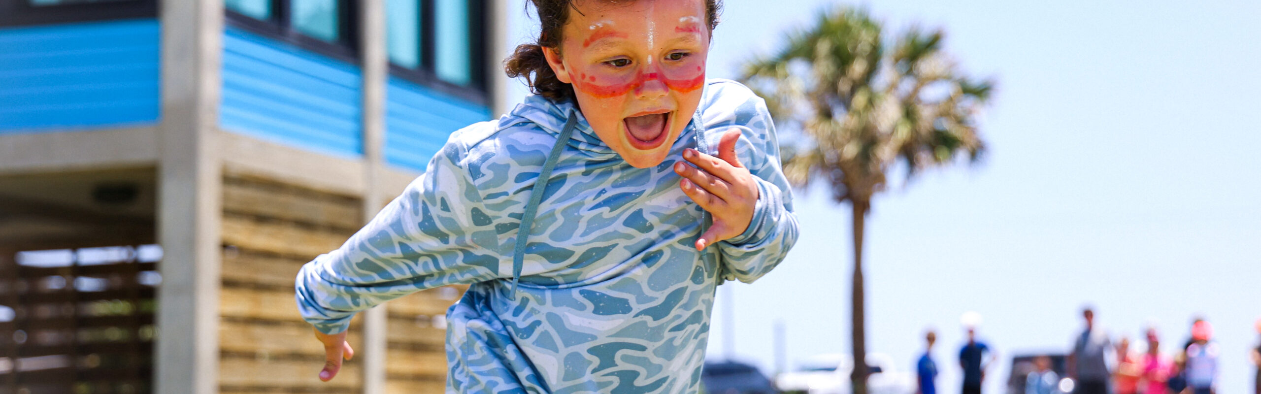 A young child with curly hair and orange face paint excitedly runs outdoors on grass near a blue building, with people and a palm tree visible in the sunny background.