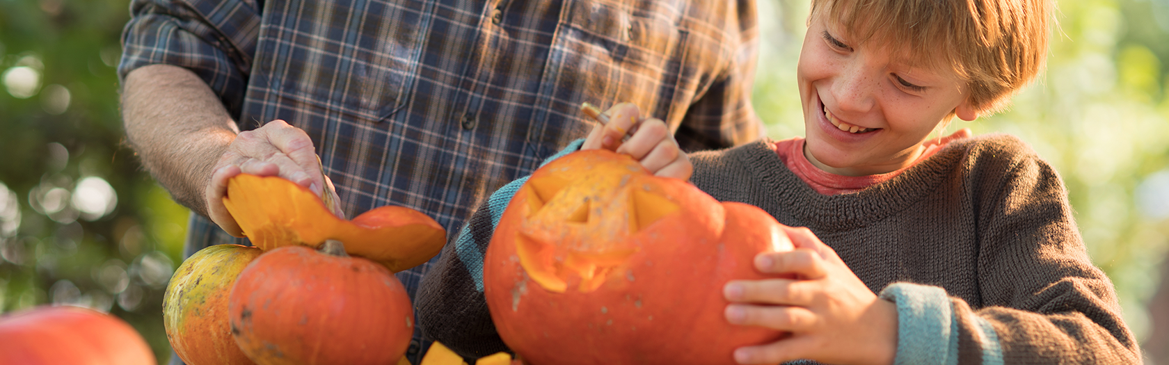 A child smiles while scooping out a pumpkin with an adult, who is holding another pumpkin. They are outdoors at a wooden table, enjoying a fall activity together.