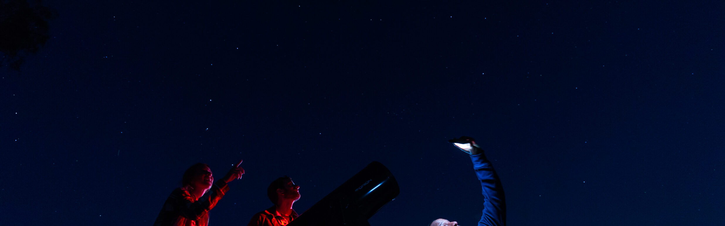Three people stargaze at night beside a large telescope; two point toward the stars while another holds up a phone or device, all under a clear, starry sky.