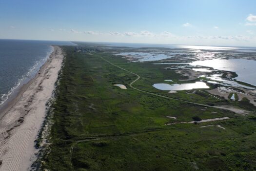 Aerial view of a long sandy beach bordered by green grassy land and scattered ponds, with the ocean on the left and wetlands on the right under a clear blue sky.