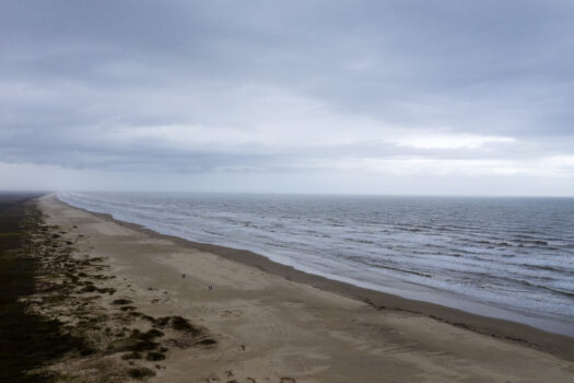 A wide, empty beach stretches into the distance under a cloudy sky, with gentle waves rolling onto the sand and sparse vegetation along the shoreline.