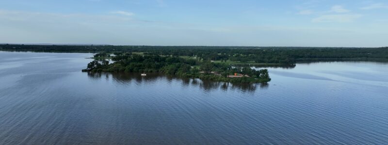 Aerial view of a green, tree landscape with open blue lake water, and distant rolling hills under a blue sky.
