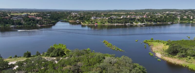 An aerial view of a blue lake with a boat traveling down it and a park with green treetops under a bright blue sky. Low hills and a few buildings are visible in the distance.