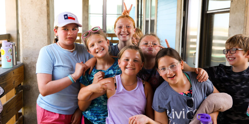 A group of smiling kids pose together on a porch, some making peace signs. They wear casual summer clothes and look happy, with sunlight streaming in from outside.