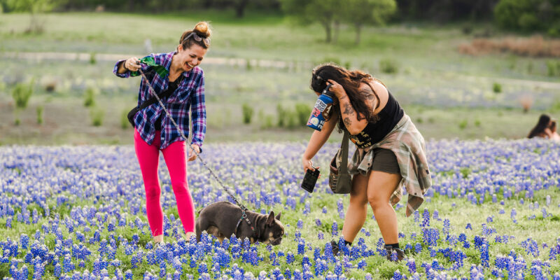 Two women laugh and walk a dog through a field of bluebonnet flowers, holding drinks. One wears bright pink leggings and a plaid shirt; the other has a tattooed arm and ties a jacket around her waist. Trees and grass fill the background.