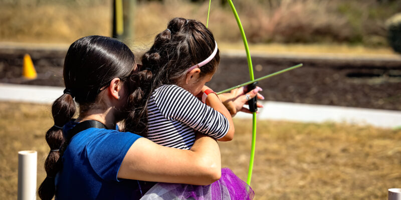An adult helps a young girl aim a green bow and arrow outdoors on a sunny day. The girl wears a striped shirt and tutu, and they are standing on dry grass with blurred greenery and a sidewalk in the background.