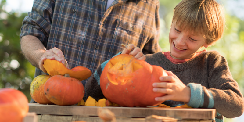 A child smiles while scooping out a pumpkin with an adult, who is holding another pumpkin. They are outdoors at a wooden table, enjoying a fall activity together.