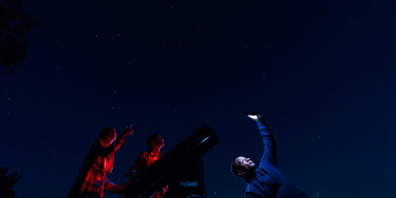 Three people stargaze at night beside a large telescope; two point toward the stars while another holds up a phone or device, all under a clear, starry sky.