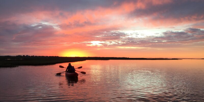 A person paddles a kayak on calm water during a vibrant sunset, with orange and pink clouds reflecting on the surface. The silhouette of the kayaker is visible against the glowing horizon.