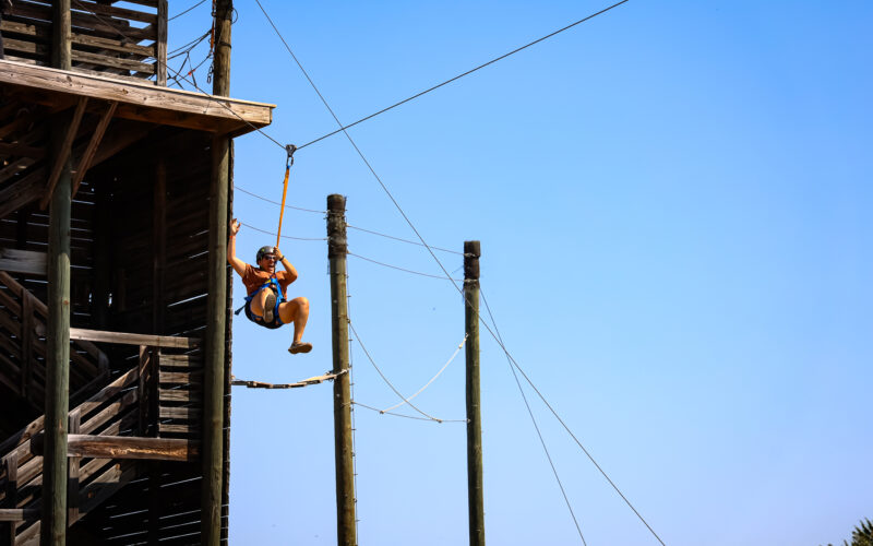 A person wearing a helmet and harness descends from a wooden platform on a zip line against a clear blue sky, with several tall utility poles and cables visible in the background.
