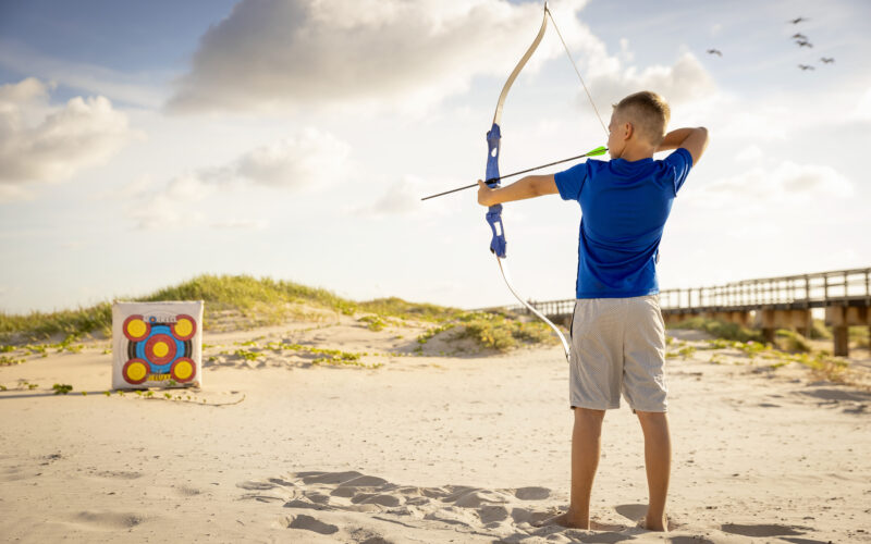 A boy in a blue shirt and gray shorts aims a bow and arrow at a colorful target on a sandy beach under a partly cloudy sky. Grassy dunes and a wooden bridge are visible in the background.