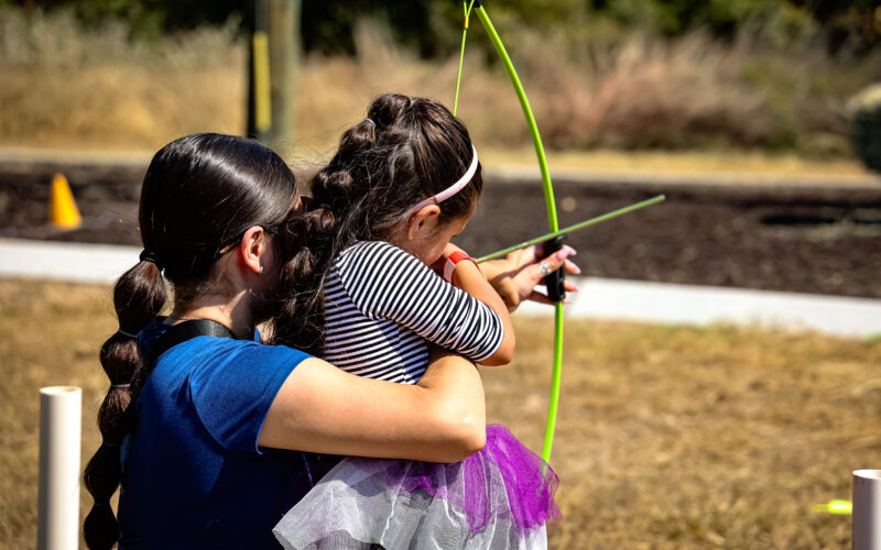 An adult helps a young girl aim a green bow and arrow outdoors on a sunny day. The girl wears a striped shirt and tutu, and they are standing on dry grass with blurred greenery and a sidewalk in the background.