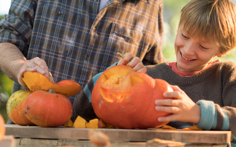 A child smiles while scooping out a pumpkin with an adult, who is holding another pumpkin. They are outdoors at a wooden table, enjoying a fall activity together.