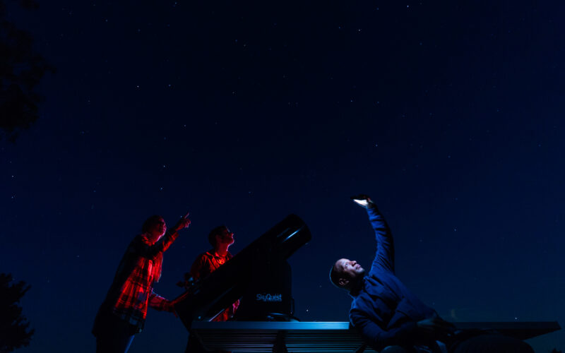 Three people stargaze at night beside a large telescope; two point toward the stars while another holds up a phone or device, all under a clear, starry sky.