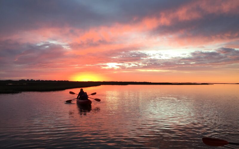 A person paddles a kayak on calm water during a vibrant sunset, with orange and pink clouds reflecting on the surface. The silhouette of the kayaker is visible against the glowing horizon.