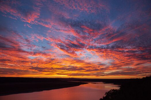 A vibrant sunset sky with dramatic orange, pink, and purple clouds reflects on a calm river, with dark silhouettes of trees and distant hills in the background.