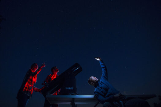 Three people stargaze at night next to a large telescope; two point at the sky, while one holds up a glowing phone, all illuminated by red and blue light under a starry sky.